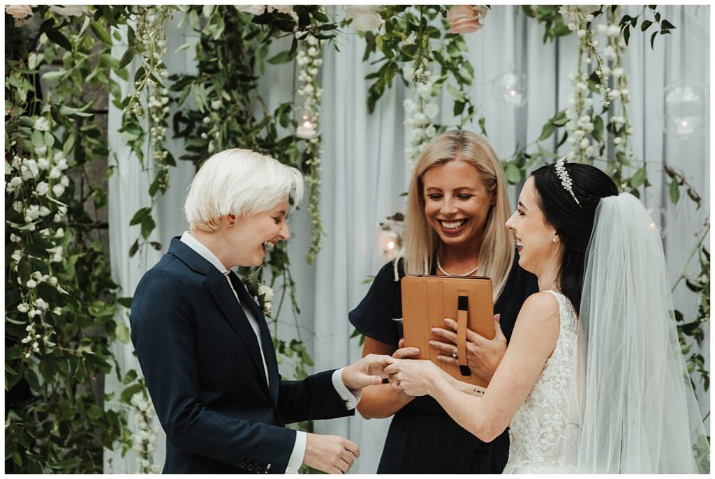 two smiling brides exchange wedding rings at floral altar during garden-themed wedding