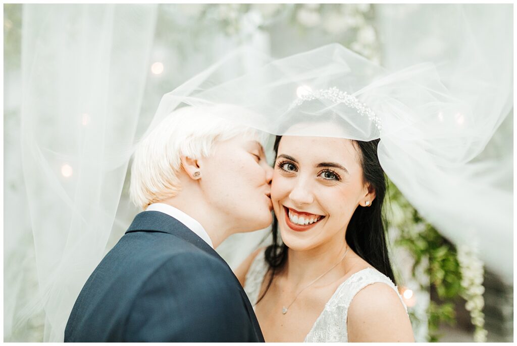 portrait of happy couple just-married sharing kiss under white veil at garden-themed wedding