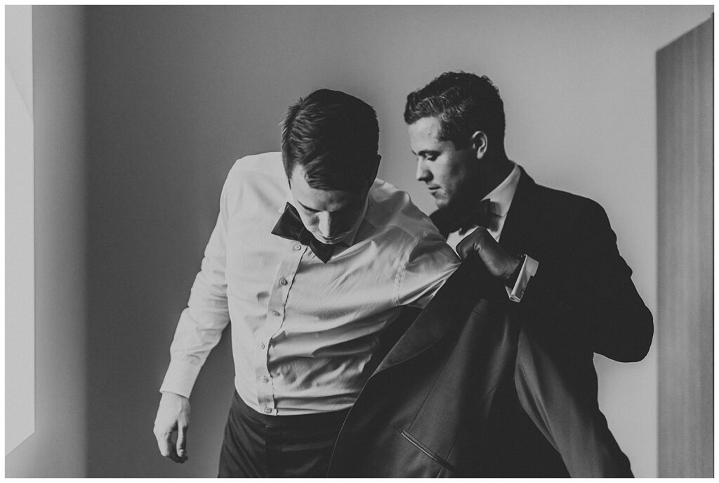 black and white photo of groom putting on black blazer with help of best man dressed in a suit