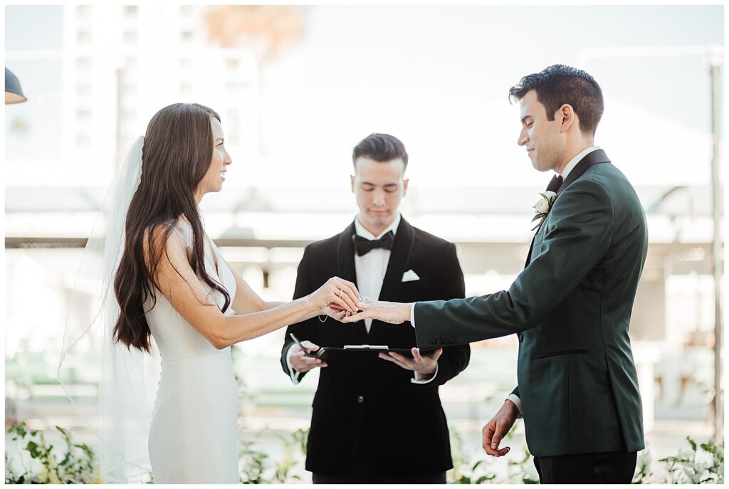 bride and groom standing in front of officiant while bride puts wedding ring on groom's finger