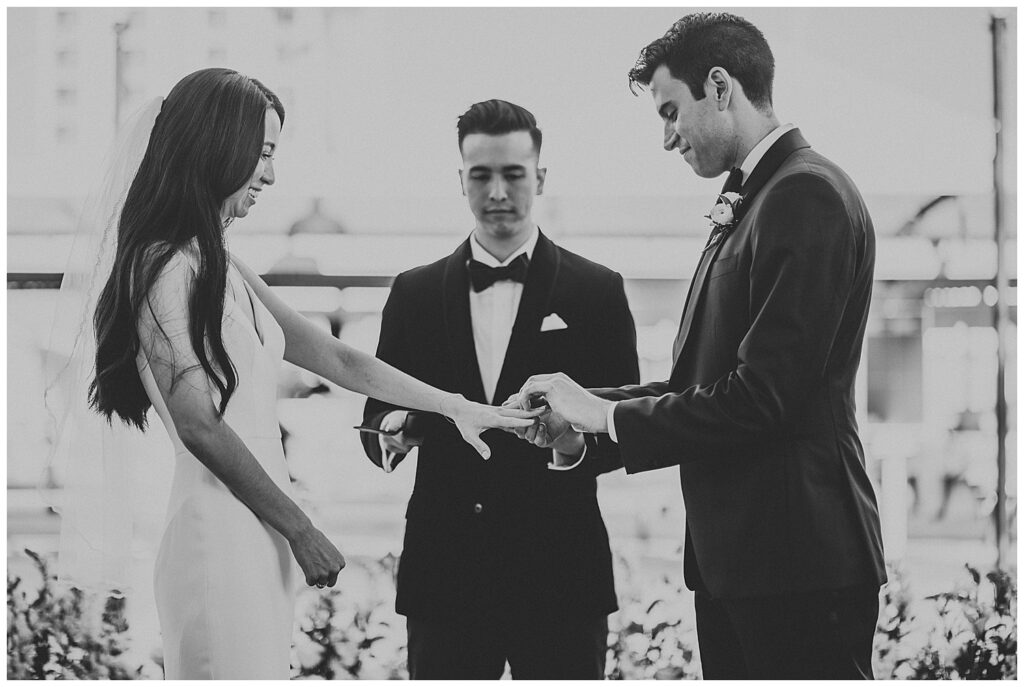 Black and white photo of bride and groom at outdoor Florida garden wedding. Groom putting wedding ring on bride during wedding day while standing in front of officiant