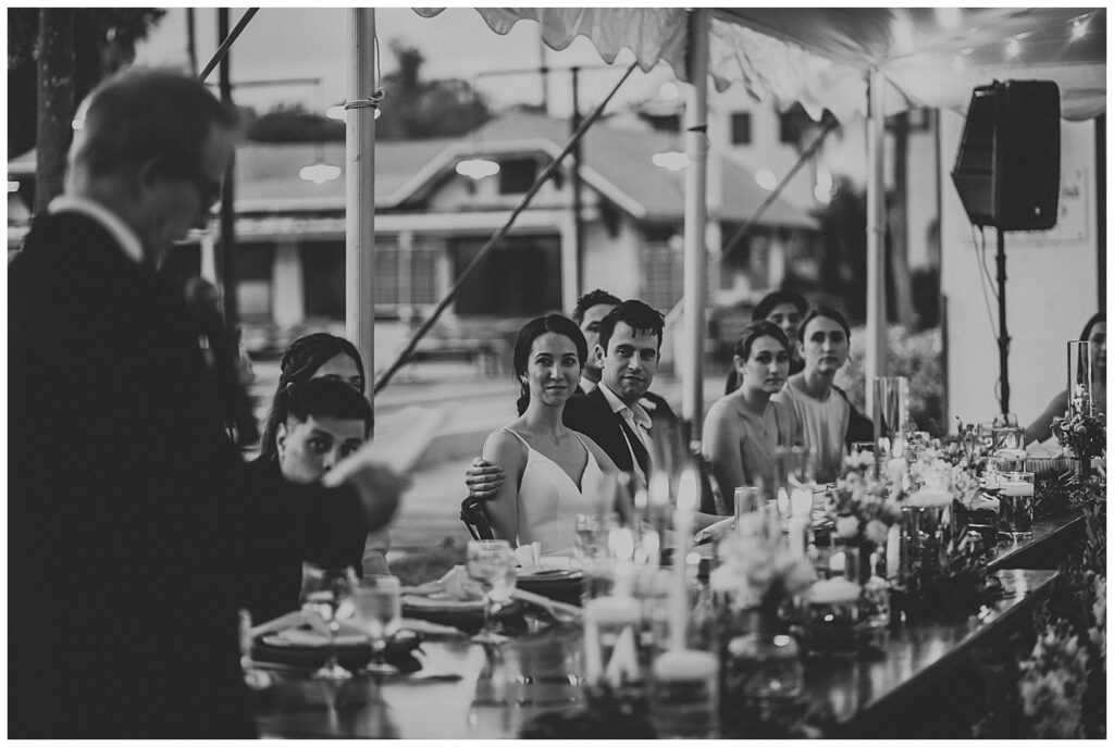 black and white photo of man giving speech while bride and groom watch smiling
