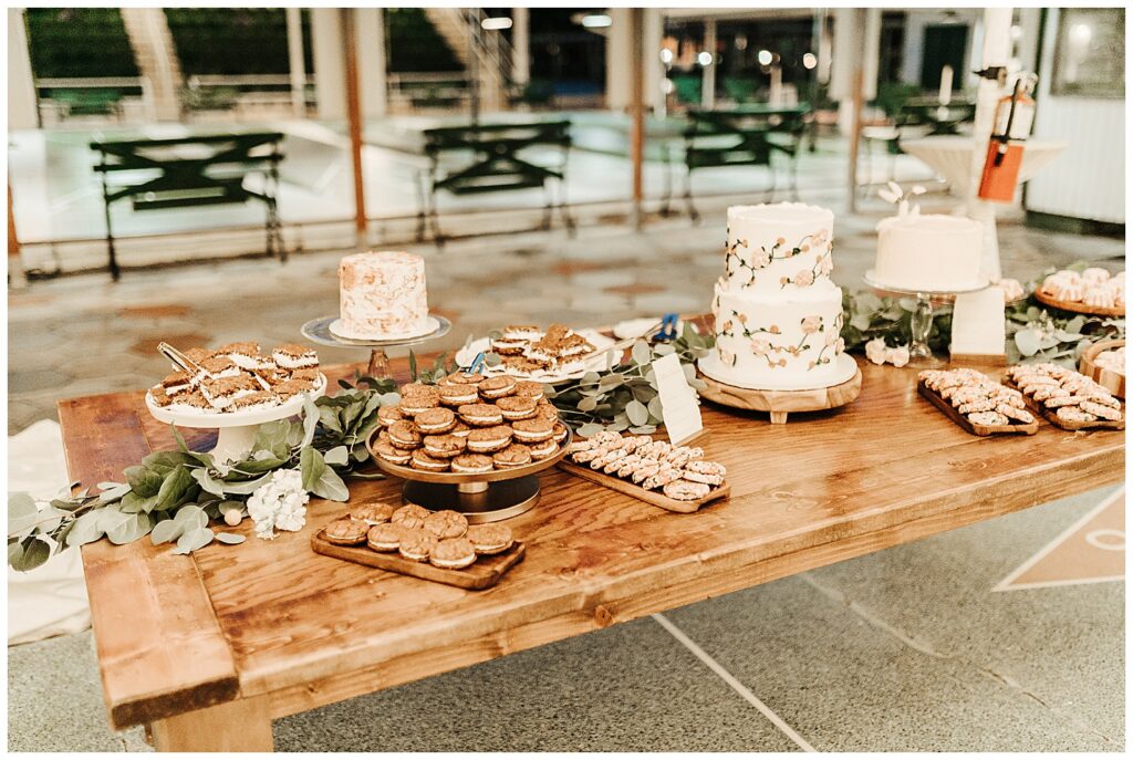 Various wedding desserts decoratively laid out on rustic wooden table lined with eucalyptus. Cookies, whoopie pies, and wedding cakes on stands