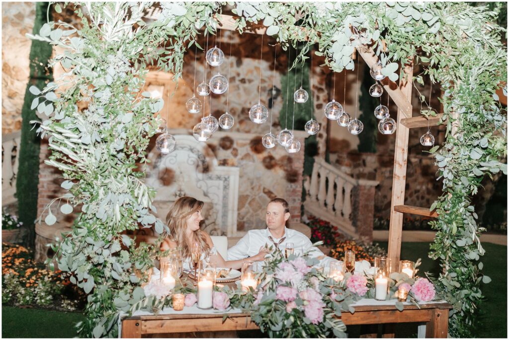 bride and groom sitting and smiling at outdoor floral wedding reception while surrounded by romantic hanging candles 