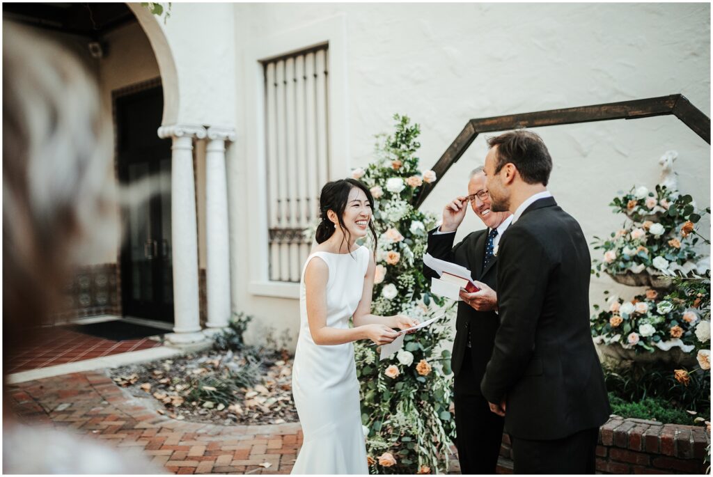 Bride and groom smiling while reading vows in front of smiling officiant surrounded by white and blush-colored roses