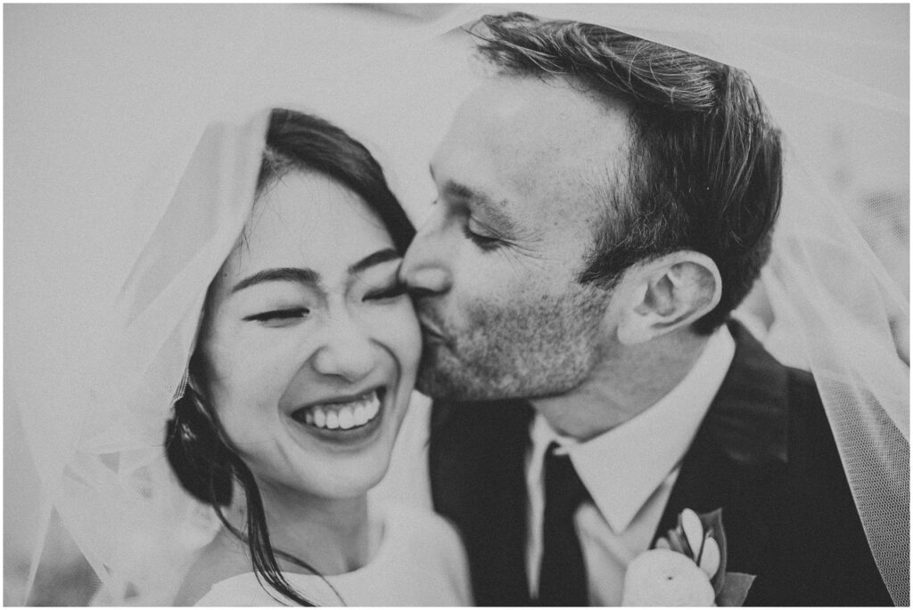 black-and-white portrait of man and woman just-married sharing kiss under wedding veil; romantic portrait of bride and groom