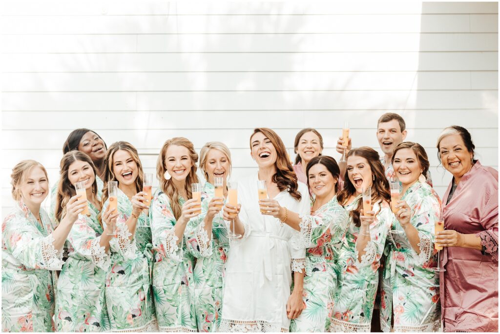 Bridal party getting ready photos; bridal party in silk robes make a toast with champagne while smiling and laughing