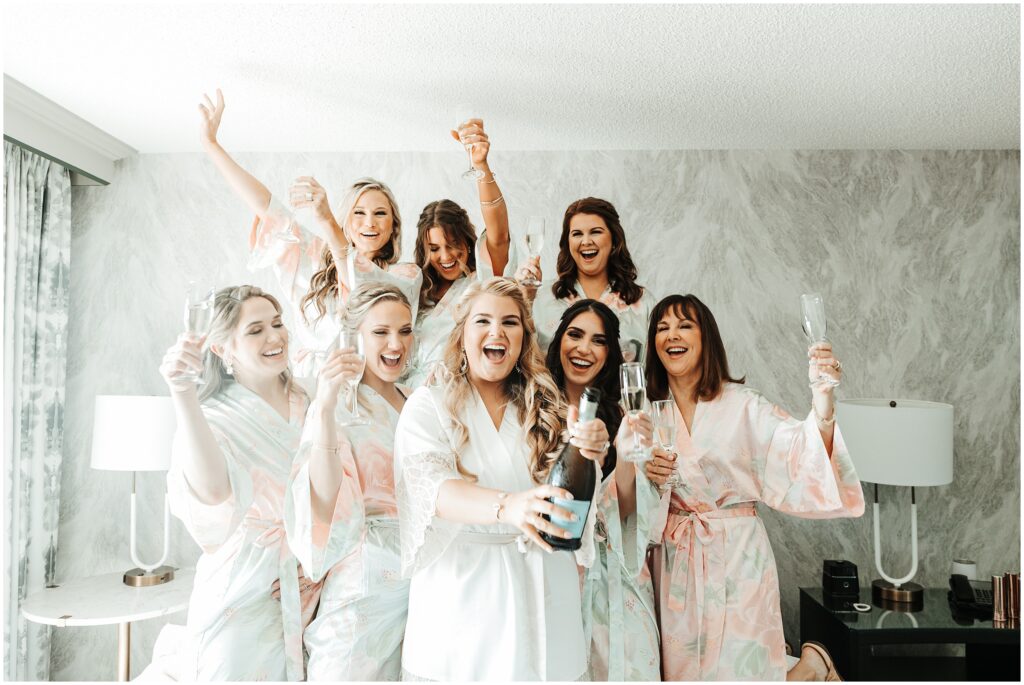 Bride and bridesmaids getting ready photo; bride in white robe pops champagne surrounded by smiling bridesmaids