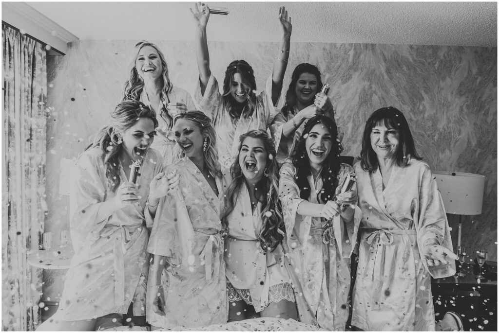 black and white photo of bride and bridesmaids wearing silk robes, smiling as confetti falls around them. Bridesmaids getting ready photos