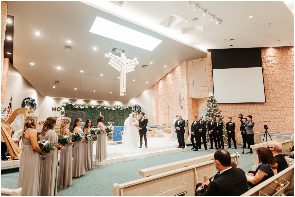 Bride and groom holding hands in front of wedding guests, surrounded by bridal party at altar 