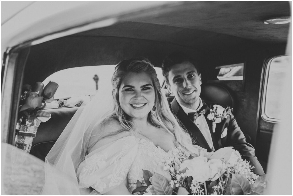 Black-and-white photo of bride and groom smiling inside of vintage white BMW car 