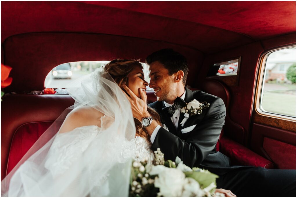 Bride and groom looking into each other's eyes as they sit on red velvet seats of a white vintage BMW car