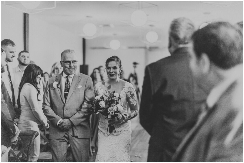 Black and white photo of bride in lace gown being walked down the aisle by father at Armature Works wedding