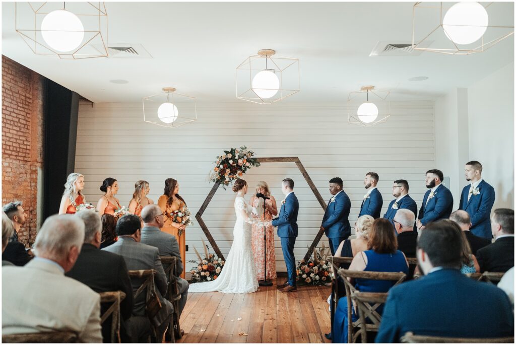 Bride and groom hold hands at altar in front of officiant, surrounded by geometric and floral details 