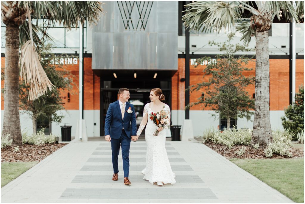 Smiling bride in white dress holds bouquet while holding hands with groom in navy suit, as they walk out of Armature Works building in Tampa FL 