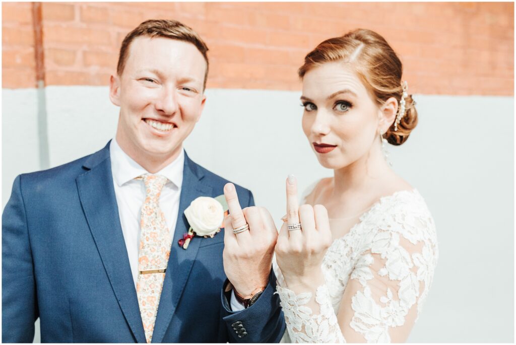 Bride and groom show off wedding bands on ring fingers in front of orange and gray wall in Downtown Tampa