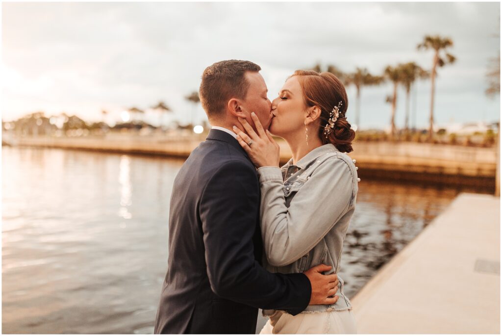 Red-haired bride in jean jacket kisses groom during sunset by the Hillsborough River after Armature Works wedding