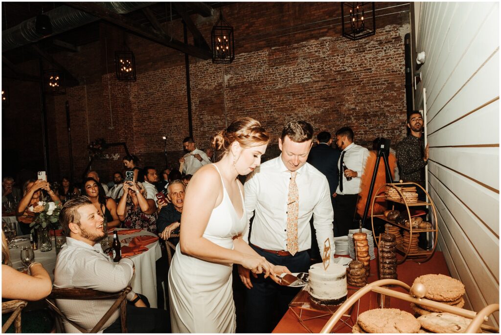 Bride and groom slice into white wedding cake in front of smiling guests at Armature Works wedding reception