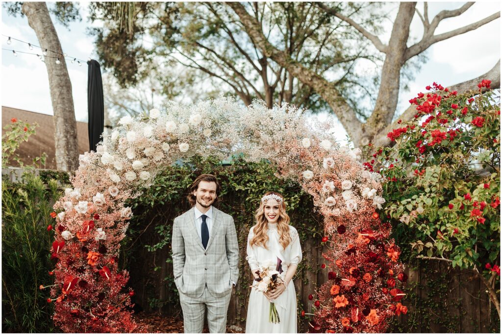 Bride and groom at intimate backyard wedding smiling under red and white gradient floral wreath
