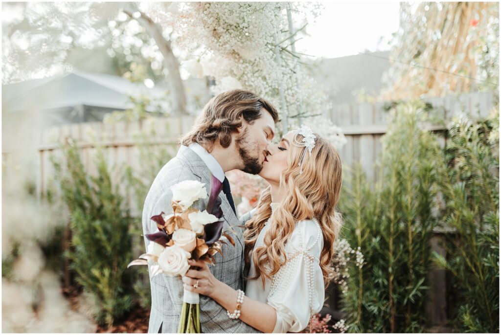 Brown-haired groom and blonde bride embrace and kiss while holding bouquet during intimate backyard wedding in Tampa, FL 