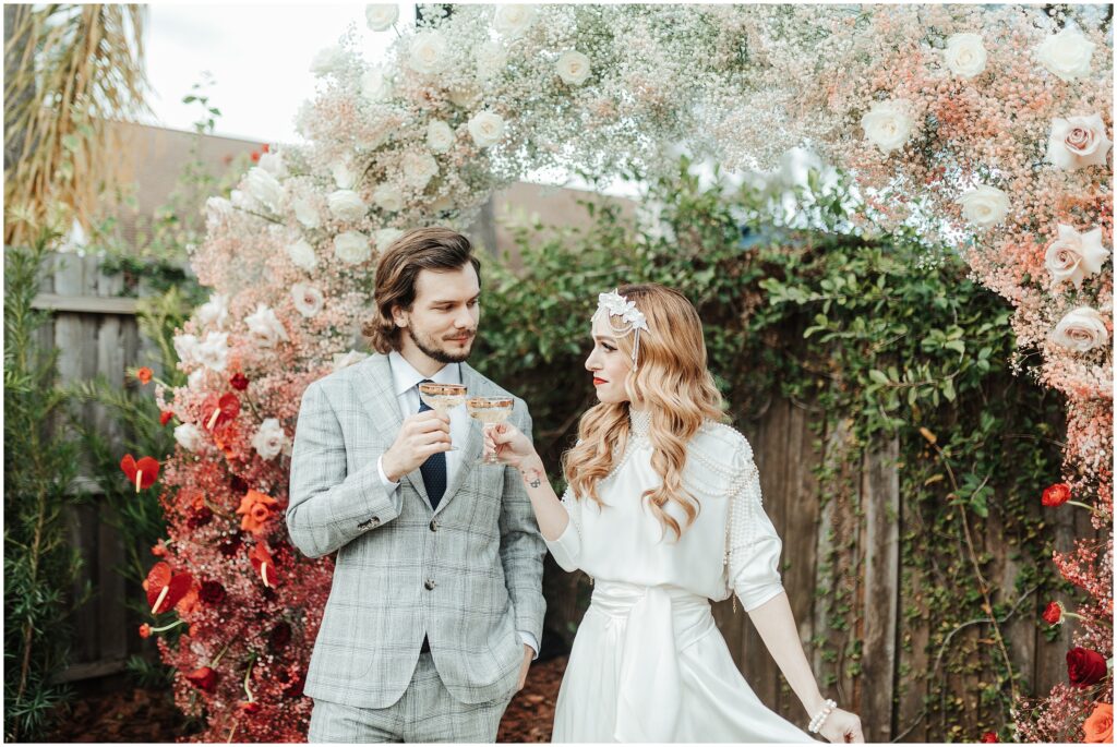 Blonde bride in white dress and brown-haired groom in grey plaid suit toast champagne under floral wreath during wedding in Florida