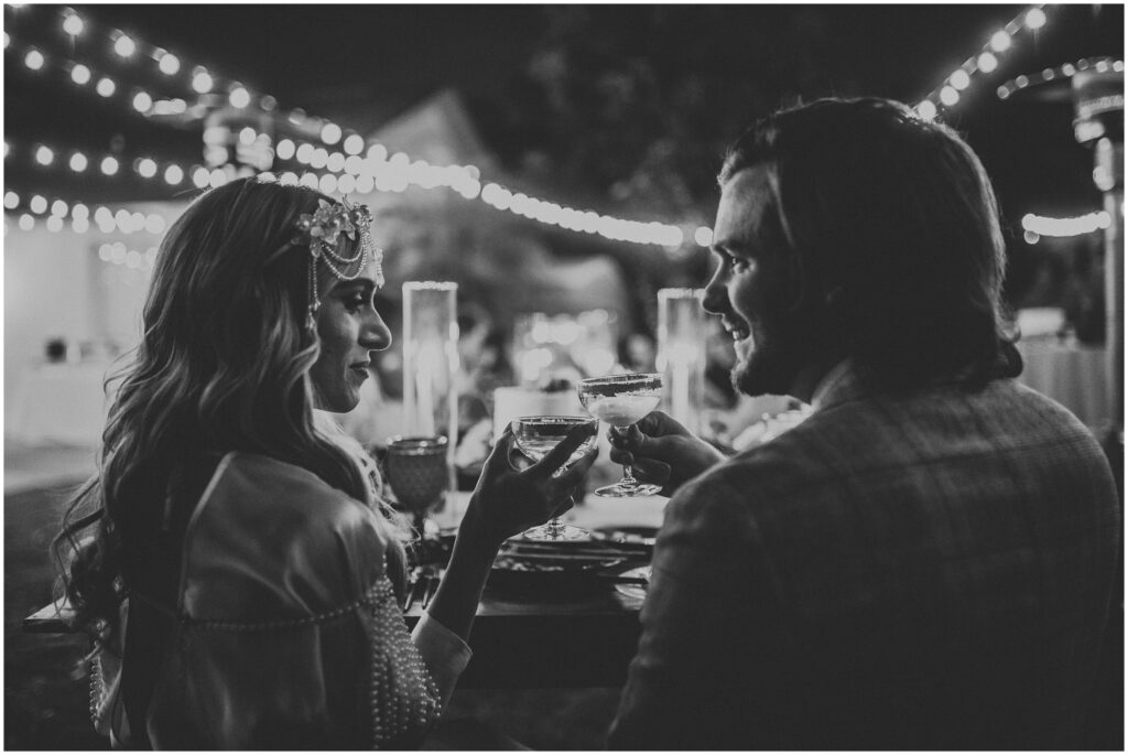 Close-up black and white photo; bride and groom toast champagne under romantic hanging lights in Tampa FL backyard wedding 