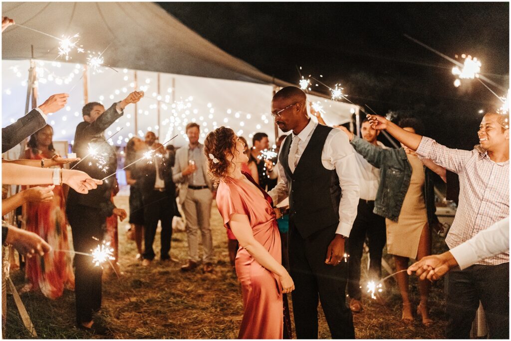 Bride and groom embrace as wedding guests wave sparklers around them 
