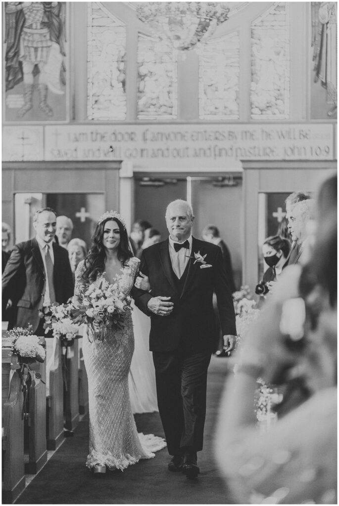 Father in black tuxedo walks smiling brunette bride in beaded gown down the aisle on wedding day