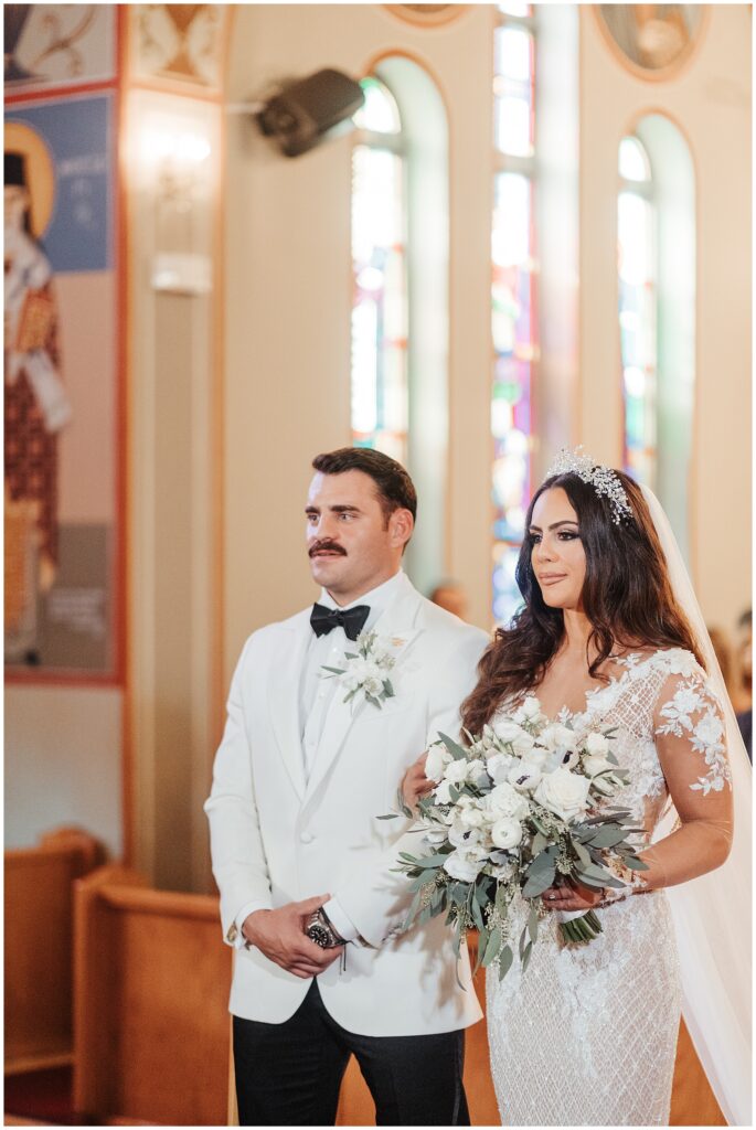 Portrait of bride and groom in all-white standing arm in arm at the altar during Greek Orthodox wedding