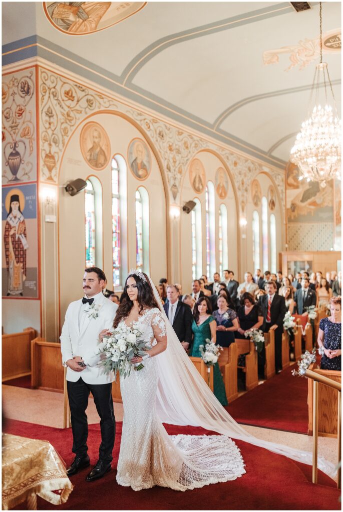 Bride and groom dressed in all white stand arm in arm at altar of Greek Orthodox church with rows of wedding guests behind them