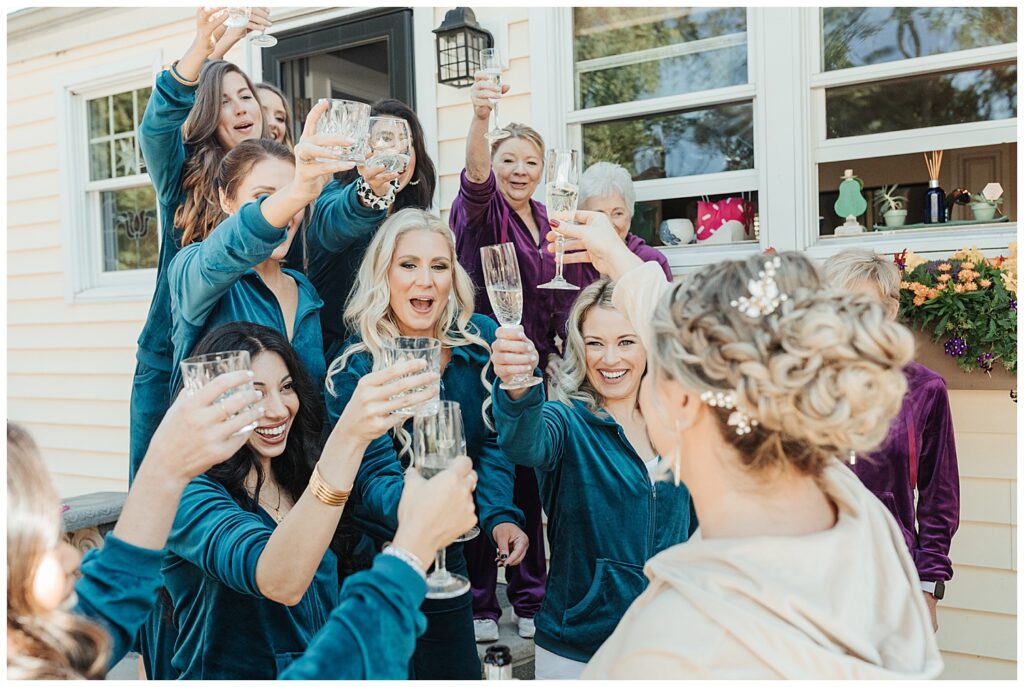bride and bridesmaids in matching jewel toned tracksuits cheer with champagne while getting ready for Crane Estate wedding