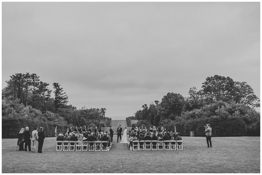 Black and white image of Crane Estate wedding ceremony; rows of chairs on open field with lush trees in background 