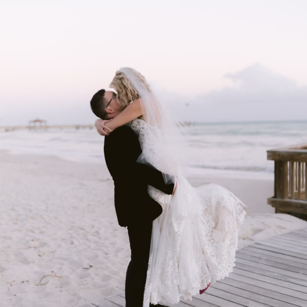Groom lifts bride in white lace wedding dress on wooden boardwalk at Tampa Bay beach wedding photographed by Ashley Izquierdo Photography