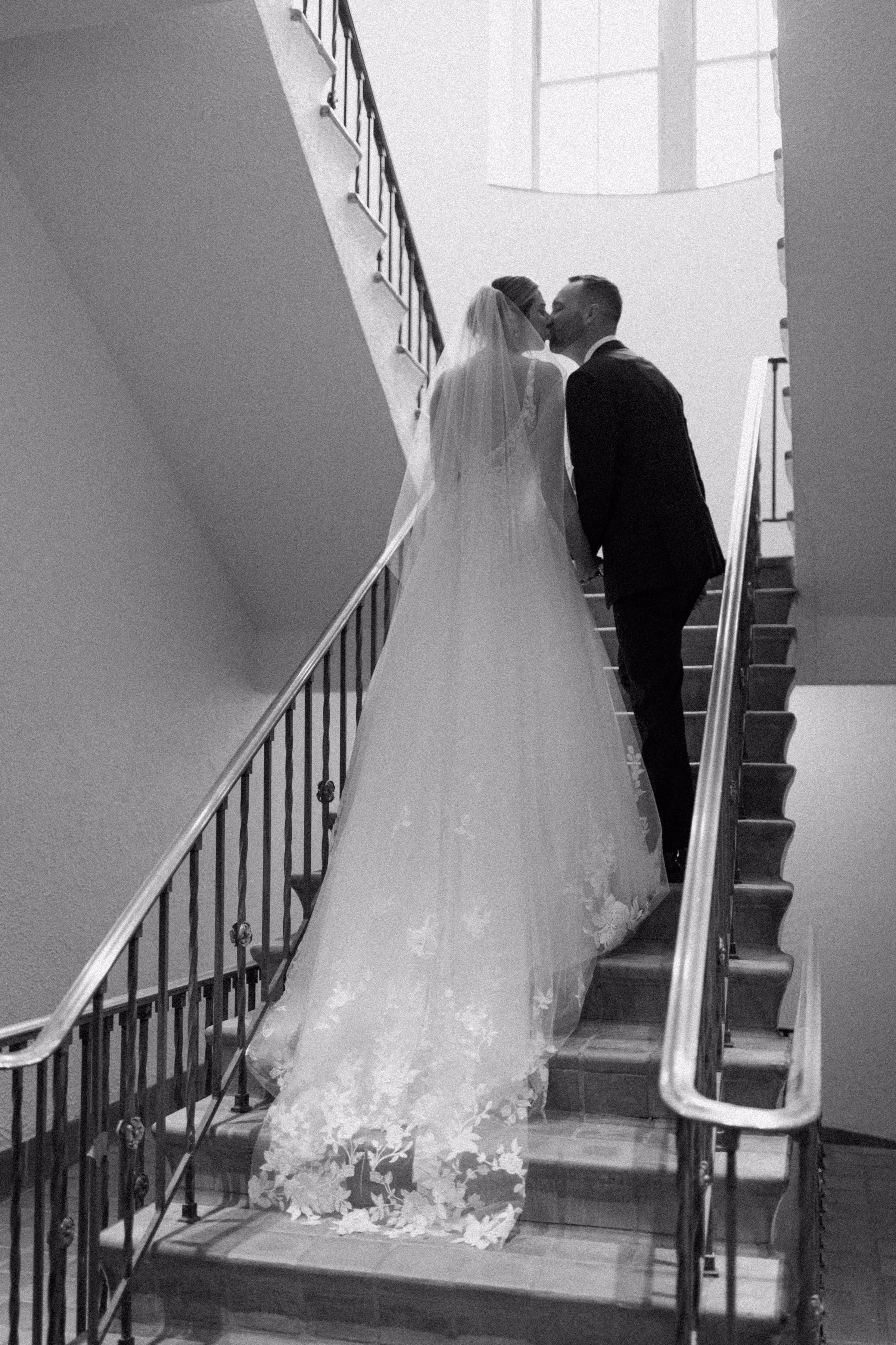 Bride and groom sharing romantic kiss on elegant curved staircase in black and white wedding photography by Ashley Izquierdo Photography in Tampa Bay