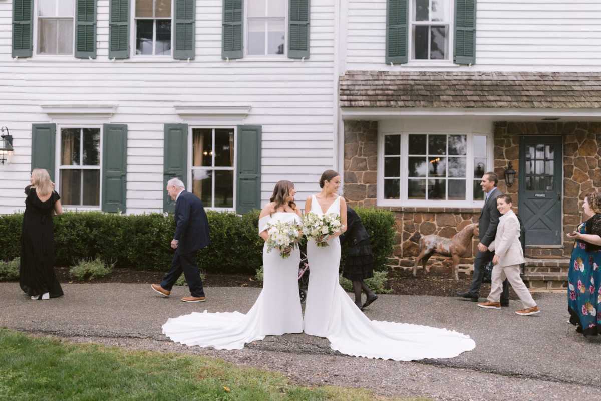 Two brides in white wedding gowns walking together during their wedding ceremony processional at a historic colonial style venue
