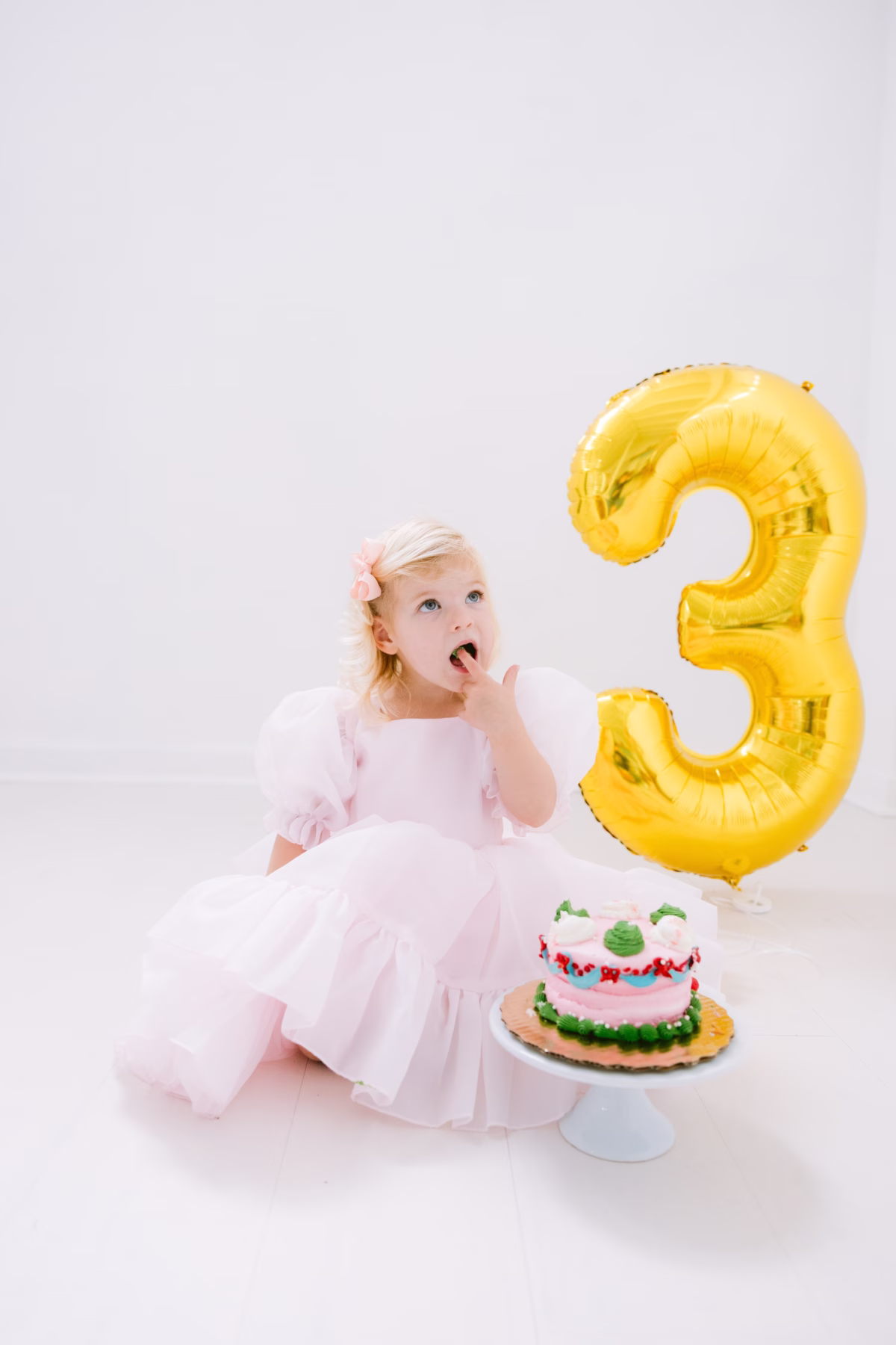 Three year old girl in pink dress celebrating birthday with cake and number three balloon during Tampa family photography session by Ashley Izquierdo Photography