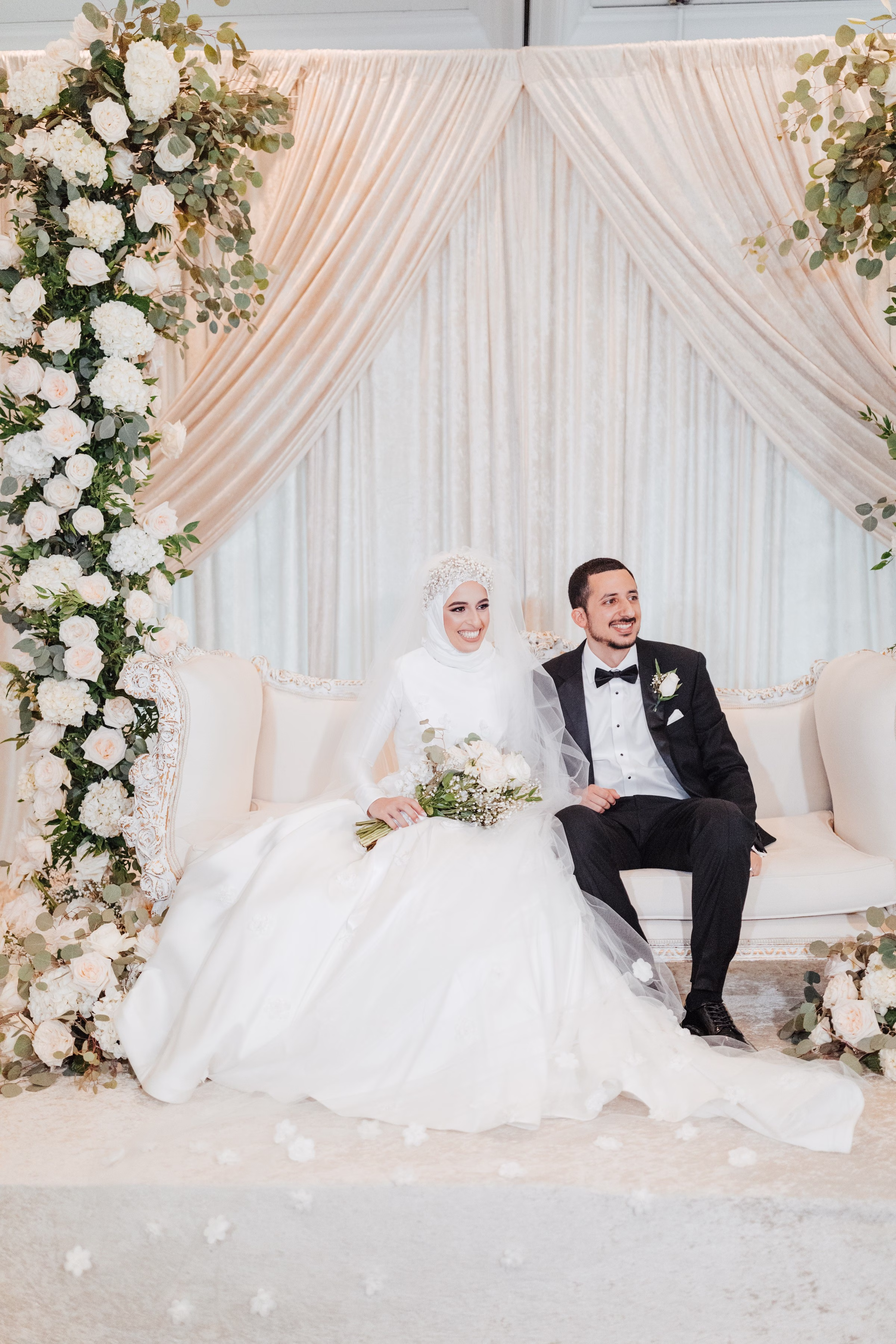 muslim bride groom white floral backdrop tampa wedding - Home Muslim bride in white hijab and groom in black tuxedo sitting on white sofa with elegant white floral backdrop at Tampa Bay wedding photographed by Ashley Izquierdo Photography