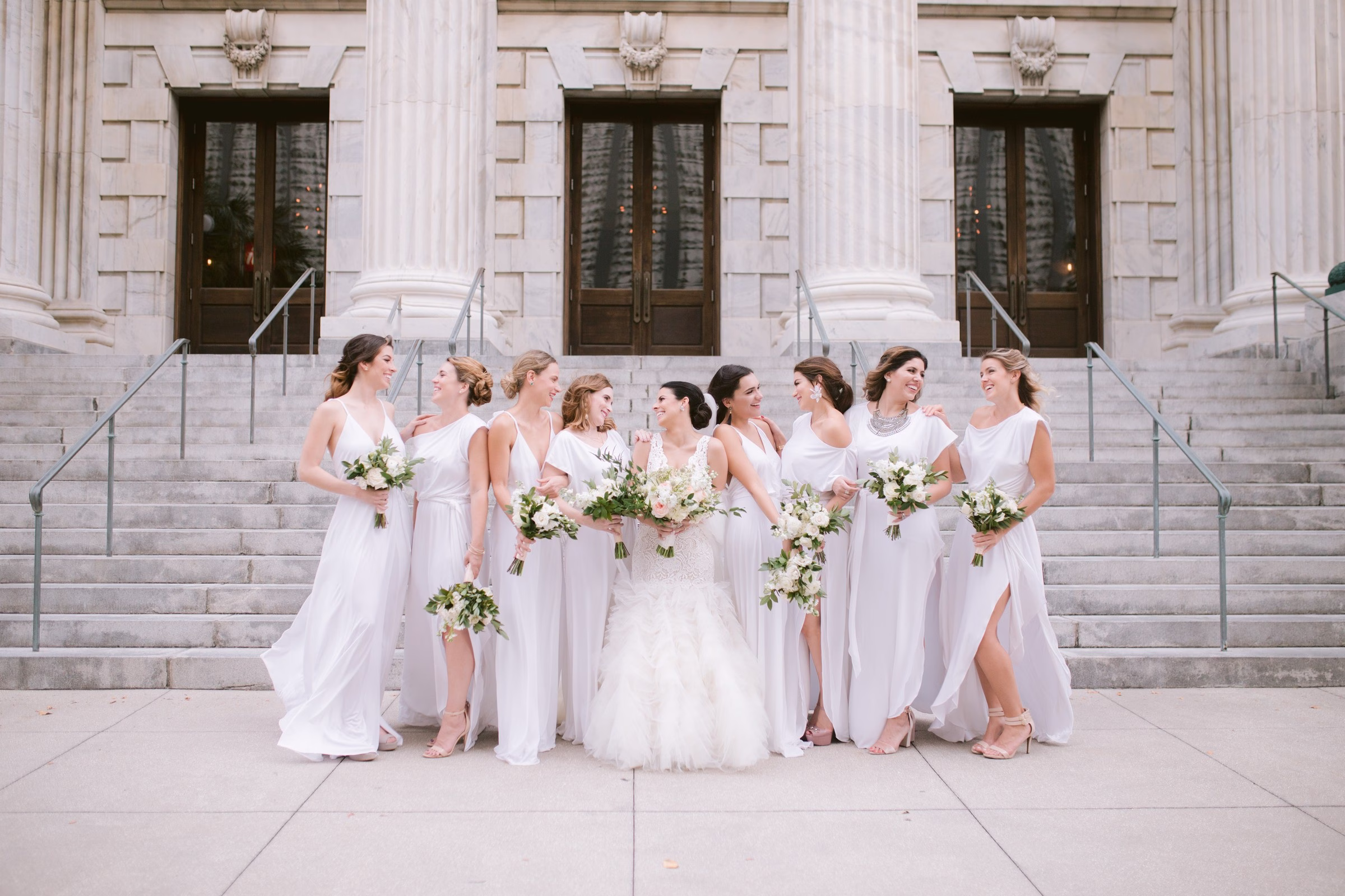 Bride and bridesmaids in elegant white dresses holding bouquets pose on the steps of Tampa Bay Museum of Art captured by Ashley Izquierdo Photography