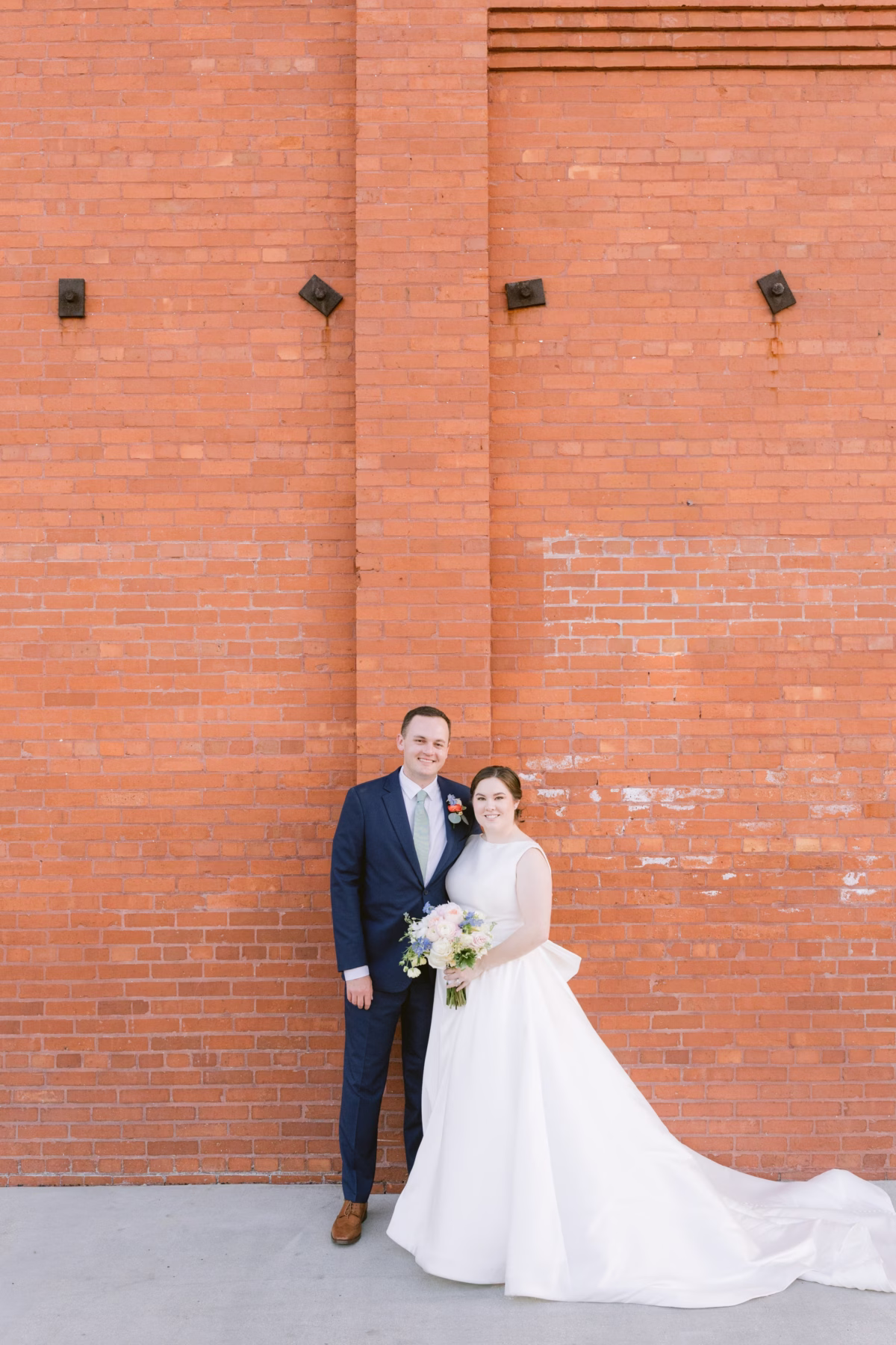 Bride and groom portrait against brick wall at Armature Works Tampa wedding featuring White Closet Bridal gown and I Dew Events florals