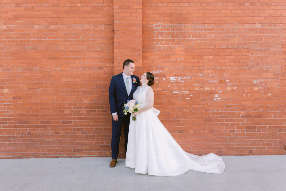 Bride in The White Closet Bridal gown with I Dew Events bouquet and groom pose against brick wall at Armature Works Tampa wedding photographed by Ashley Izquierdo Photography