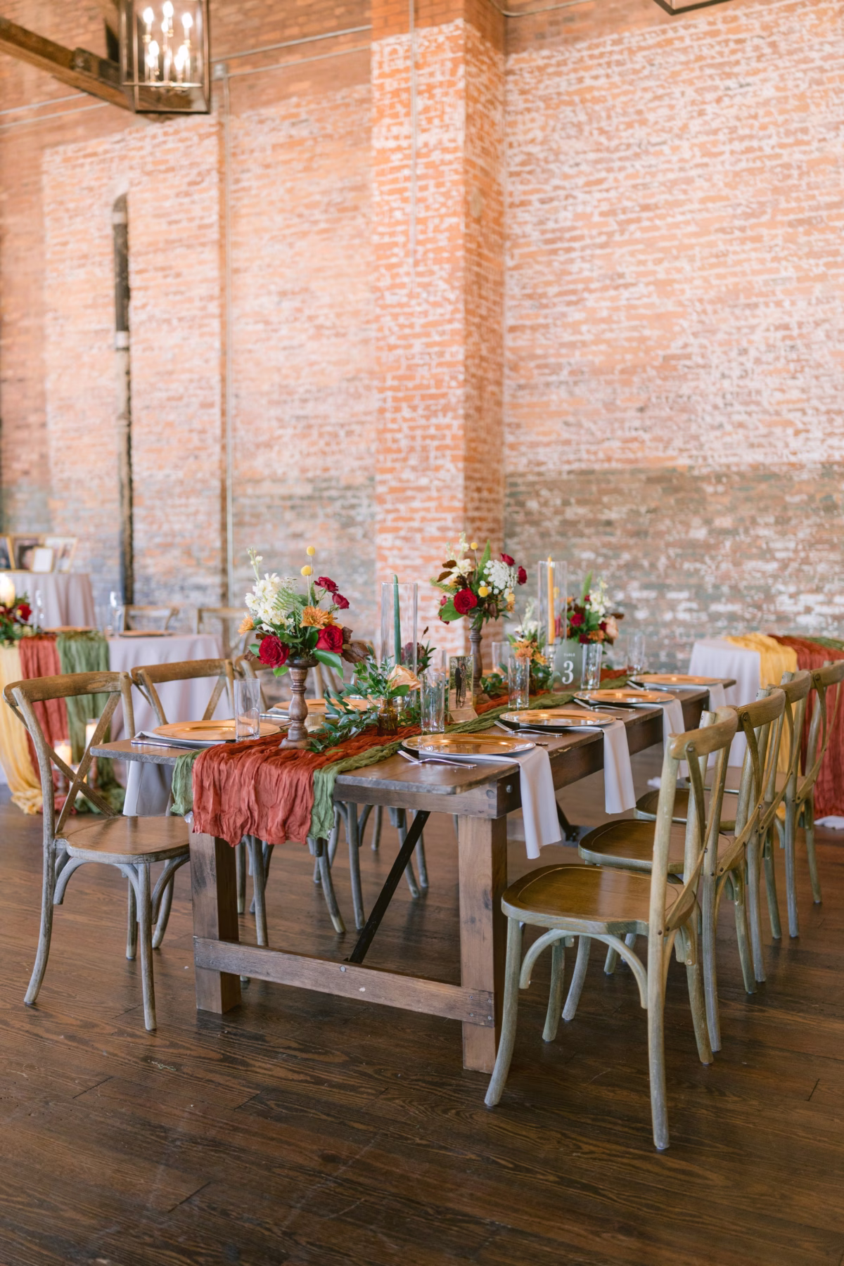 Rustic wedding reception tablescape with burgundy and sage linens, wooden farm table, cross-back chairs, and red floral centerpieces by Events in Bloom at Armature Works Tampa