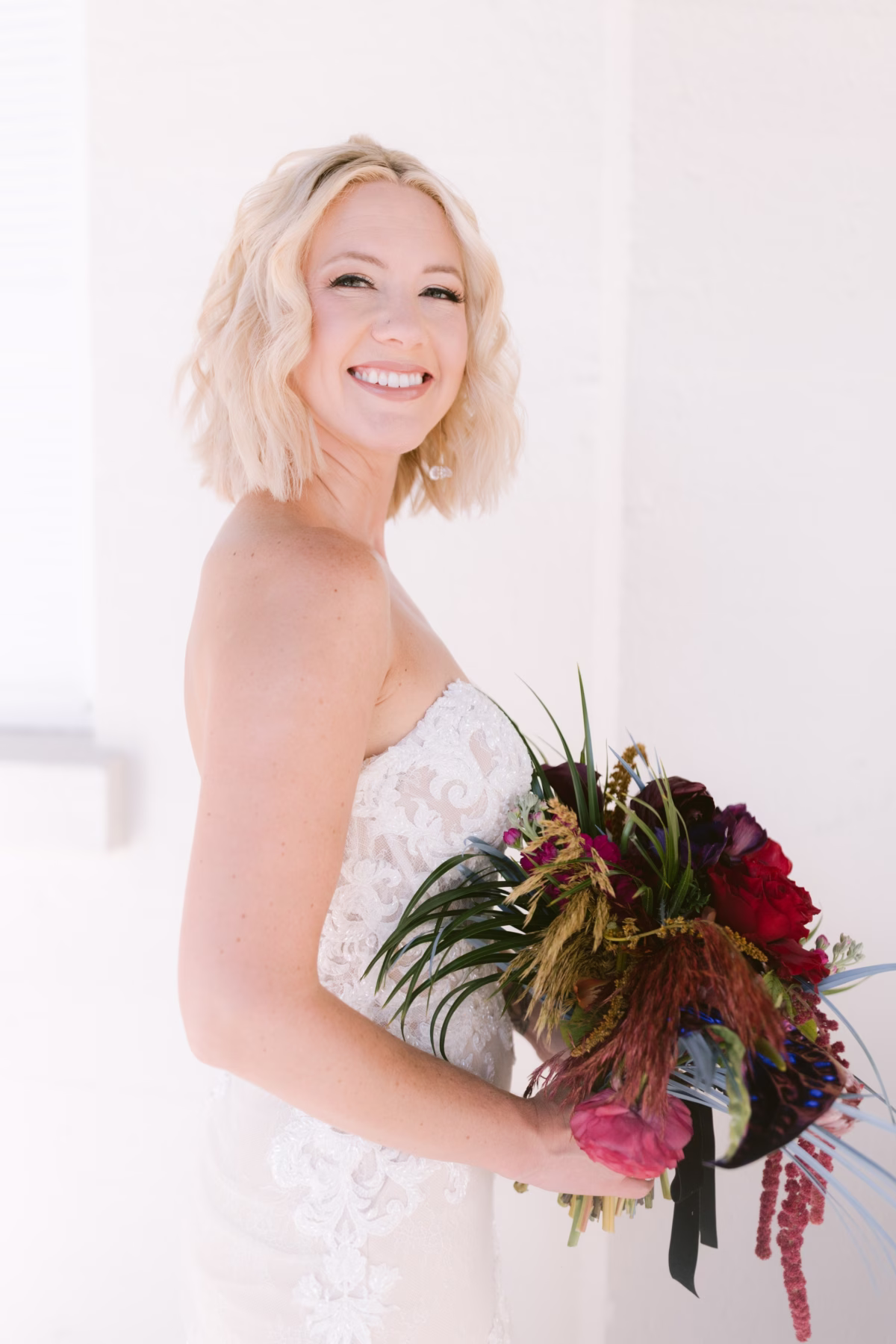 Smiling bride in white lace wedding dress holding vibrant jewel tone bouquet by Arms of Persephone at Casa Marina Key West beach wedding