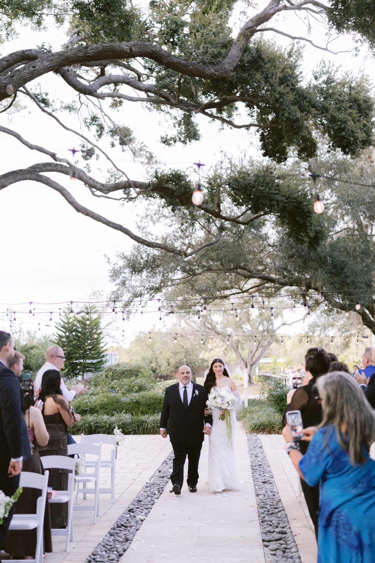 Bride walks down the aisle with her father under string lights and oak trees at Soire Estate Tampa wedding with florals by Ava Elise Decor