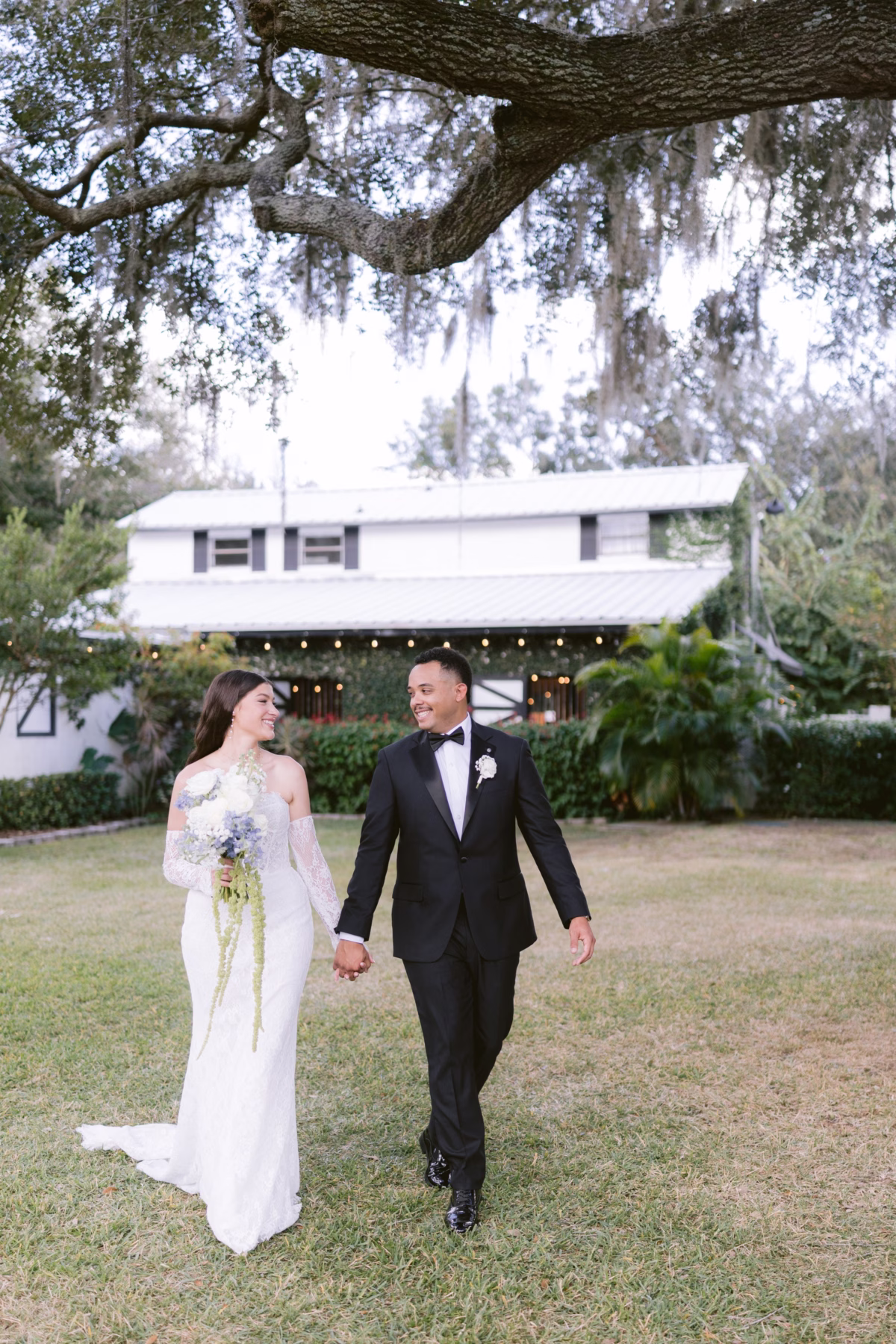 Bride in Ella Blu Bridal gown walks hand in hand with groom across lawn at Soire Estate Tampa wedding photographed by Ashley Izquierdo Photography