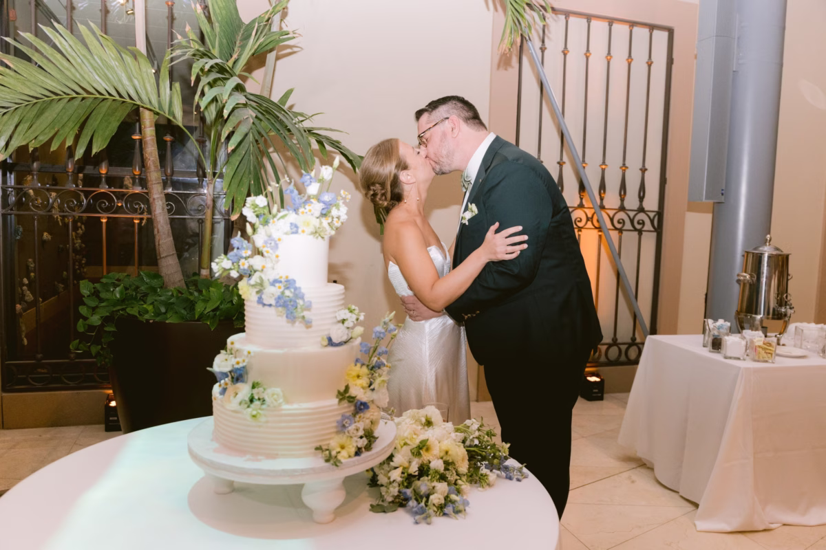 Bride and groom share a romantic kiss during cake cutting ceremony with white tiered wedding cake by Sweet Diva's Cakery at Nancy Cotto Events venue in St. Petersburg