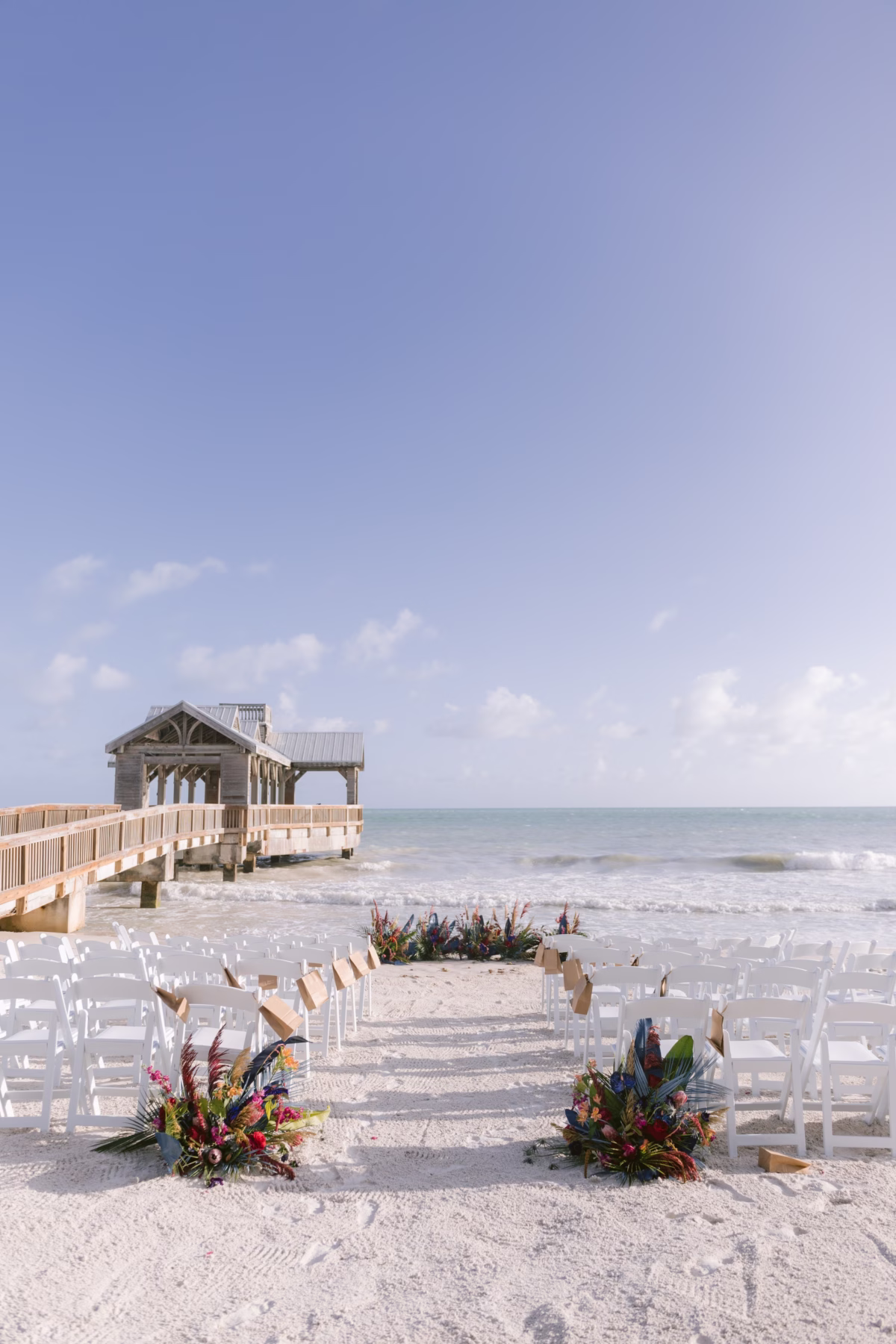 Beach wedding ceremony setup at Casa Marina Key West with tropical floral arrangements by Arms of Persephone featuring white chairs and wooden pier backdrop