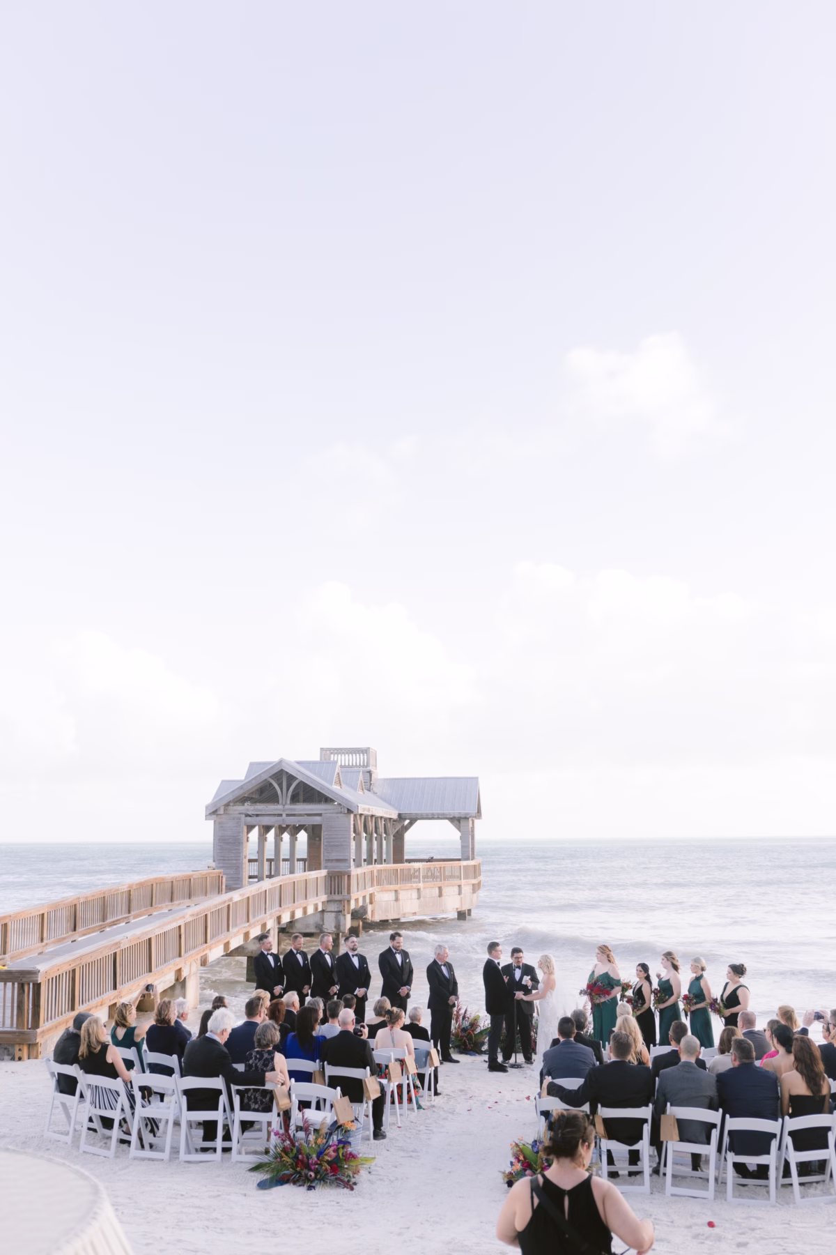 Beach wedding ceremony at Casa Marina Key West with jewel tone florals by Arms of Persephone and guests seated on white chairs facing wooden pier pavilion