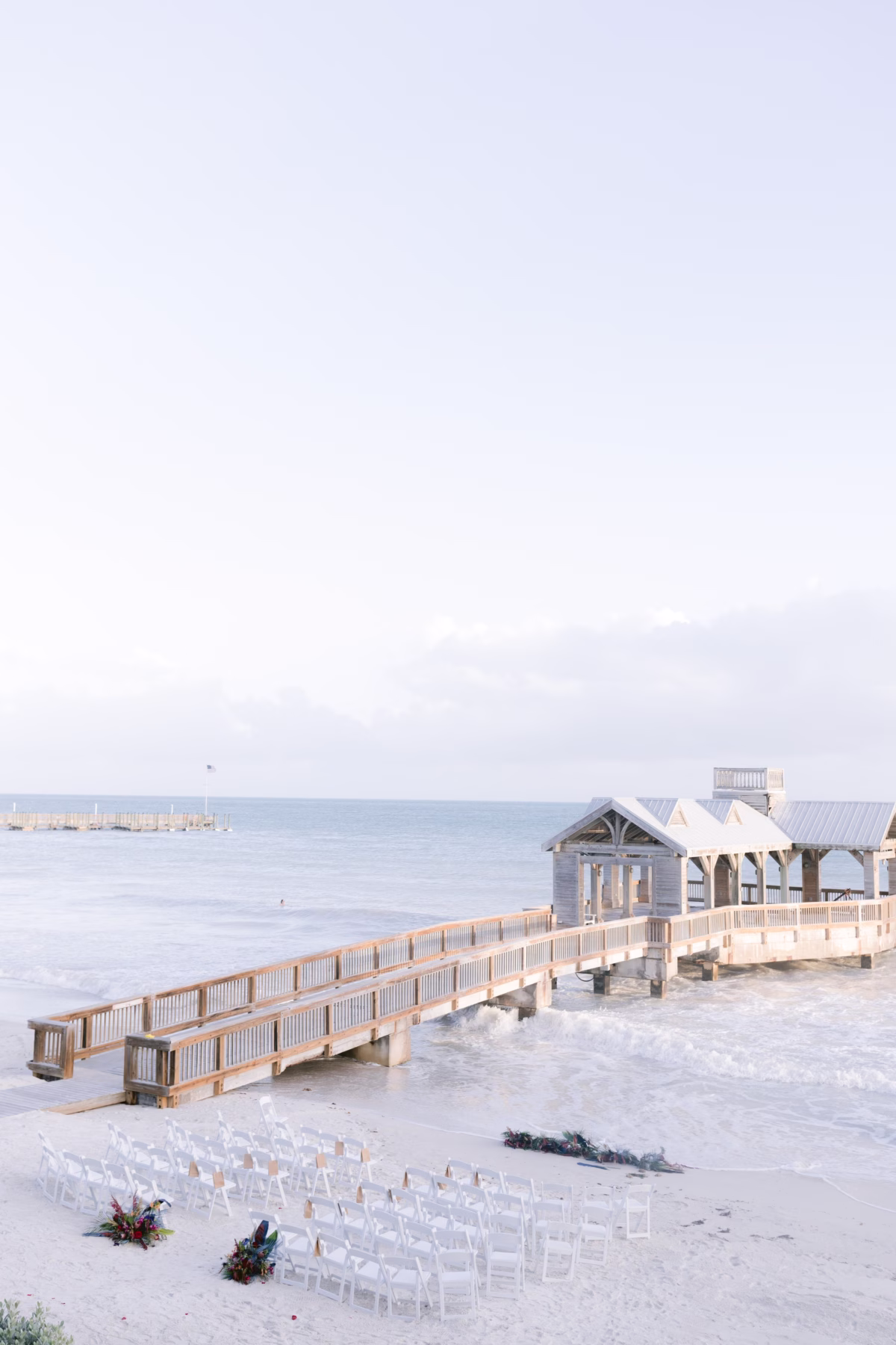 Beach wedding ceremony setup with white chairs and wooden pier at Casa Marina Key West with floral arrangements by Arms of Persephone