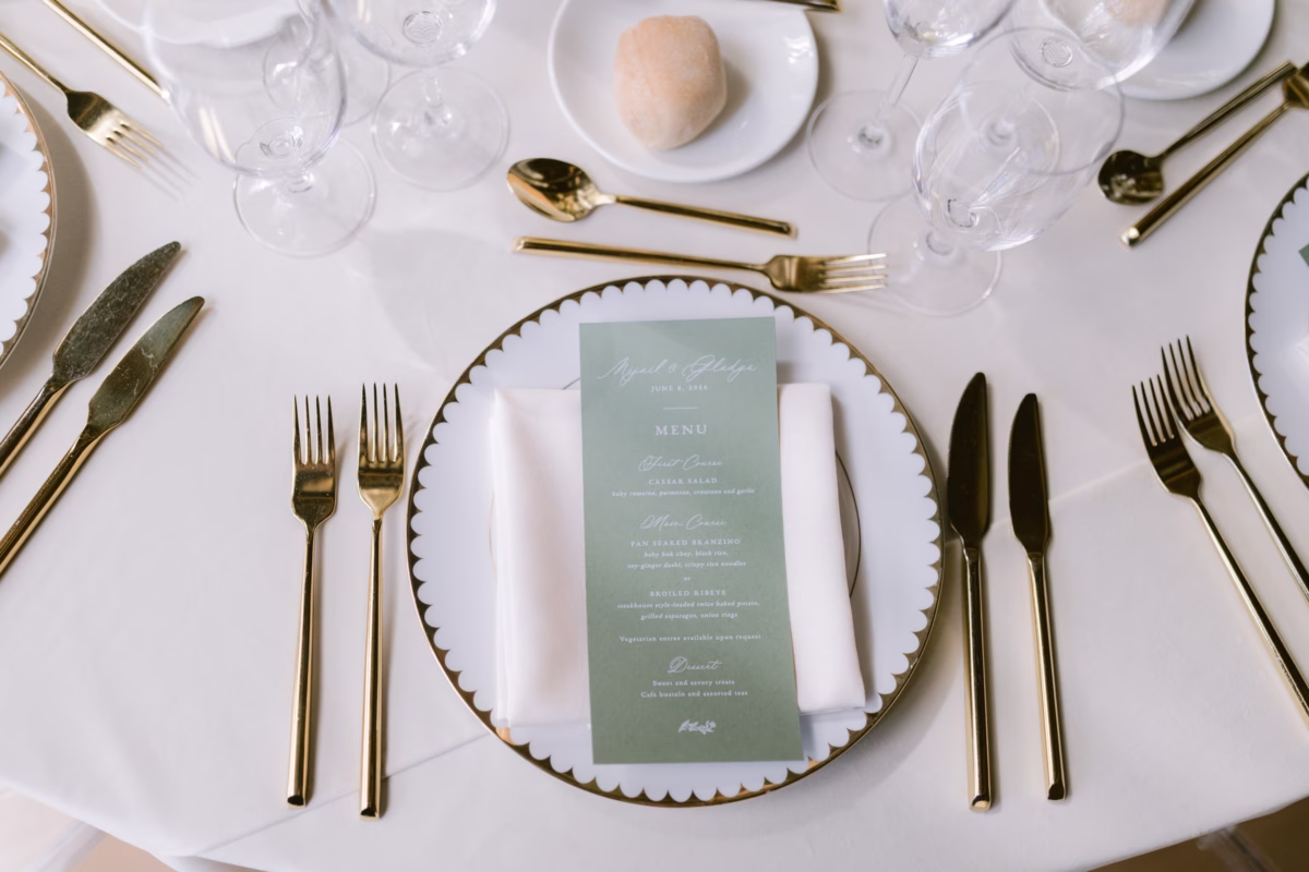 Elegant wedding table setting with sage green Reverie Press menu card, gold flatware, and scalloped charger plates at The Roundhouse Beacon in Hudson Valley