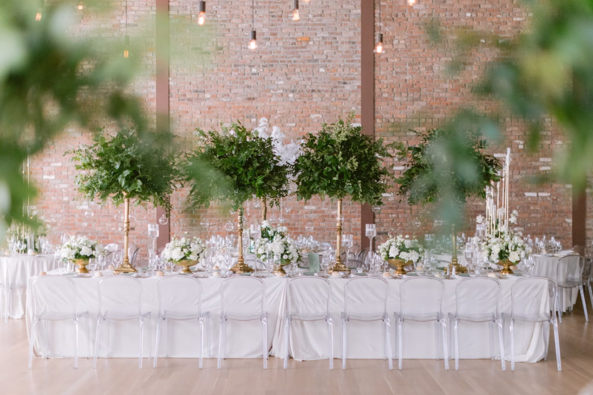 Elegant white wedding reception tablescape with tall greenery centerpieces by Maky The Diva Flowers at The Roundhouse Beacon venue featuring exposed brick walls and clear ghost chairs